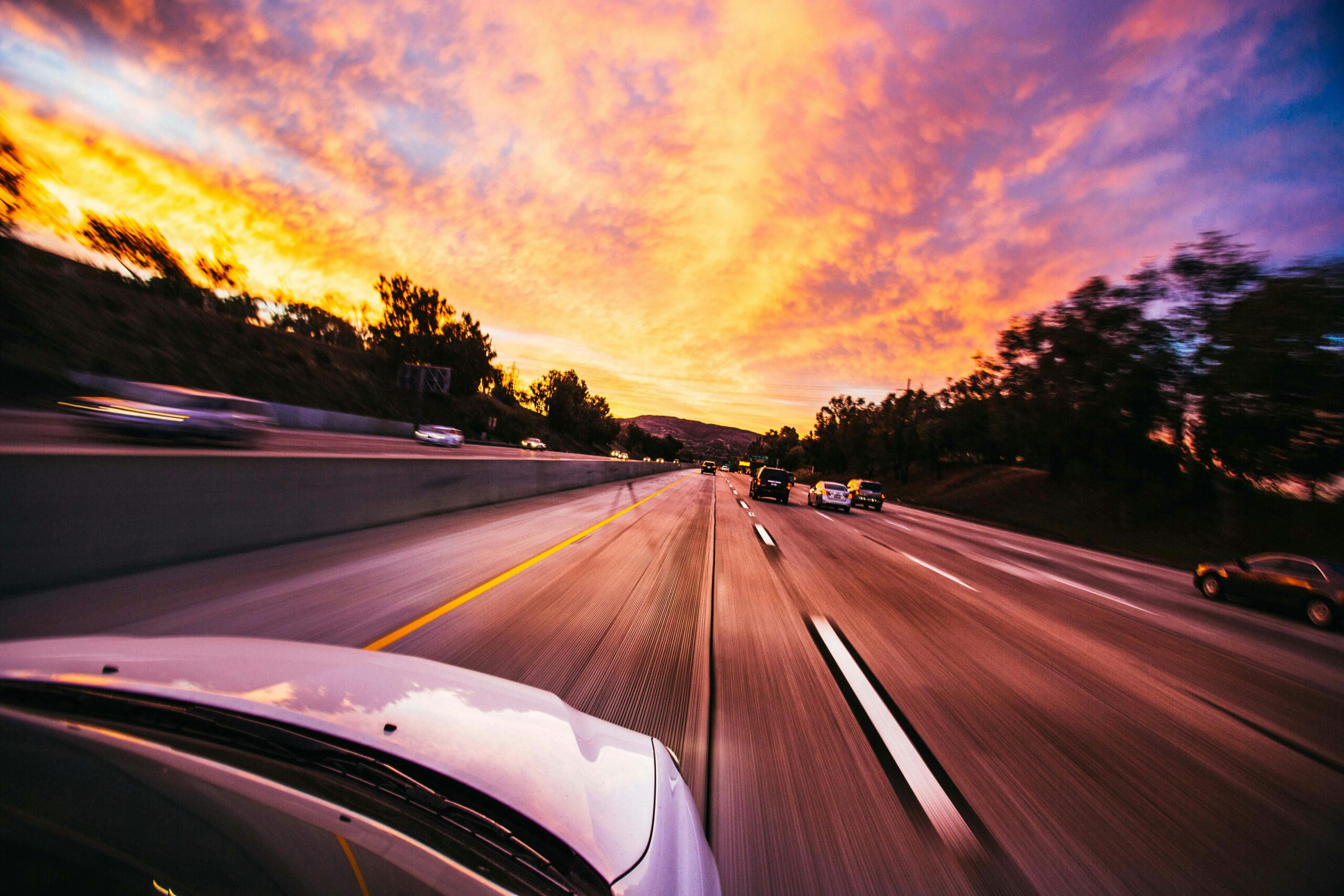 A blurred view from a moving car on a highway at sunset, showcasing vibrant clouds and taillights of vehicles, conveying a sense of speed and freedom.