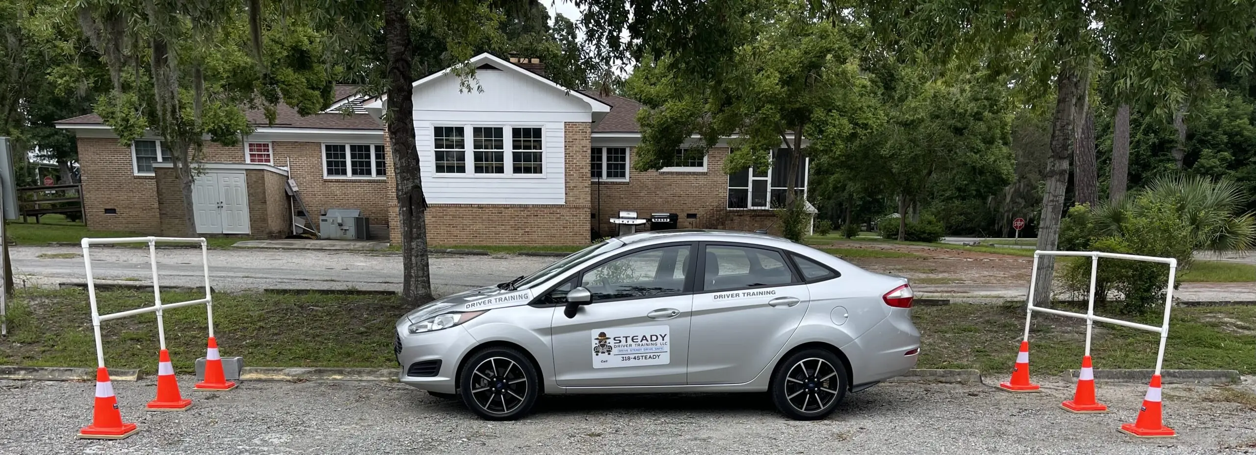 A silver car marked for driver training is parked beside a red-brick building, surrounded by traffic cones and barriers for practice.