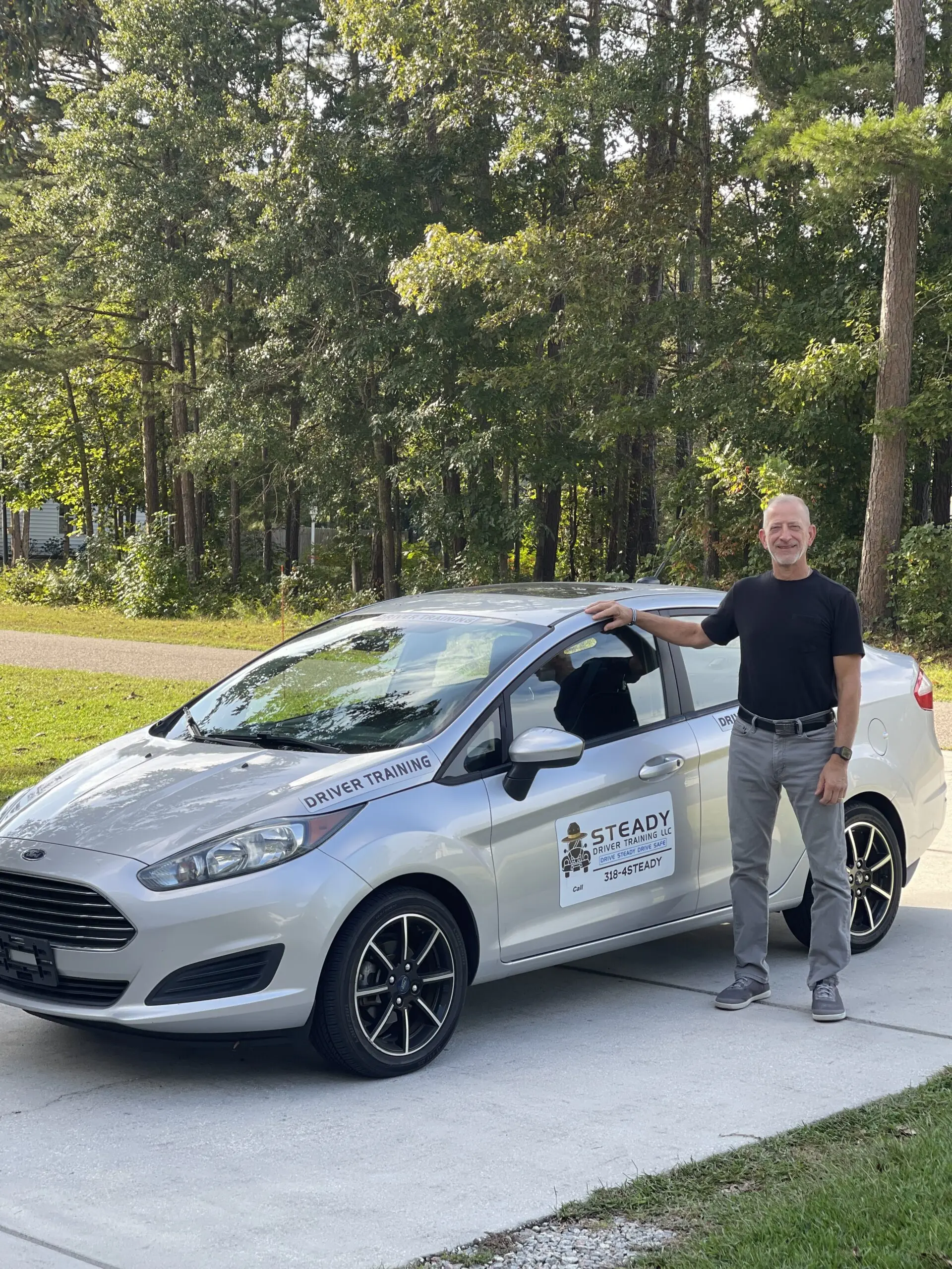 A man stands next to a silver Ford Fiesta labeled "Driver Training," set against a backdrop of trees. This image highlights driving instruction services.